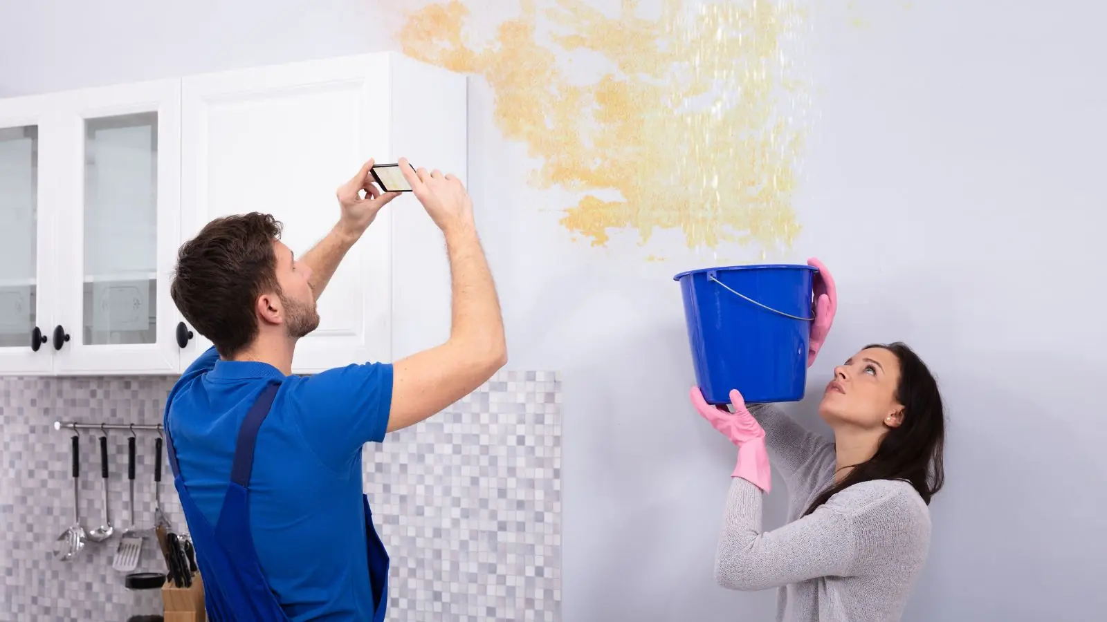 Renter photographing a water-stained wall while another person holds a bucket under the leak
