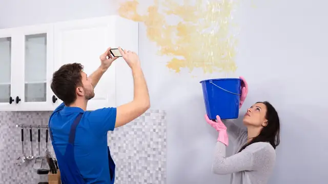 Renter photographing a water-stained wall while another person holds a bucket under the leak