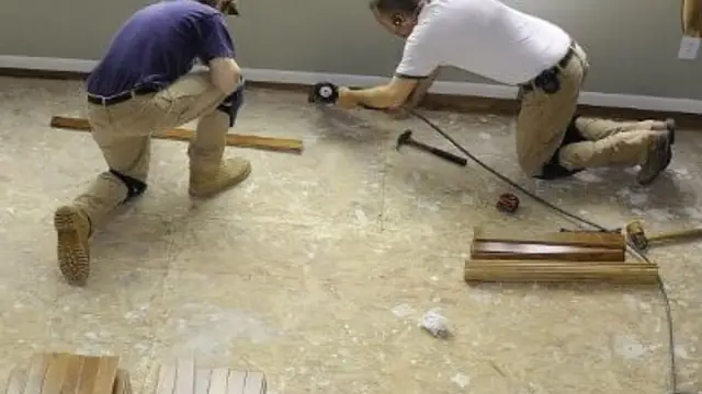 Two workers kneeling on a bare subfloor, measuring and fitting boards during a repair